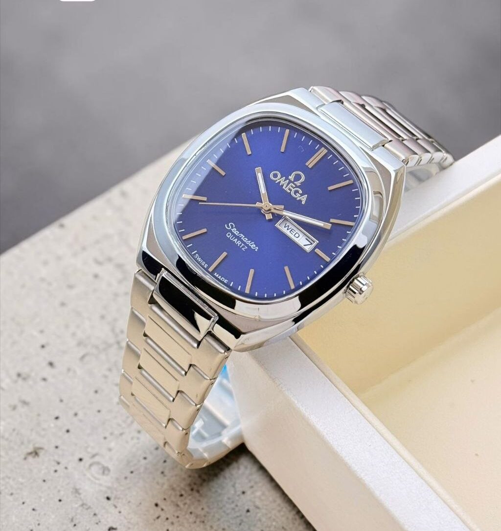 Omega men’s analog watch featuring a classic round case, stainless steel chain bracelet, working day and date display, quartz movement, and luxury-inspired styling. Omega men's analog watch featuring a classic round case, stainless steel chain bracelet, working day and date display, quartz movement, and luxury-inspired styling.
